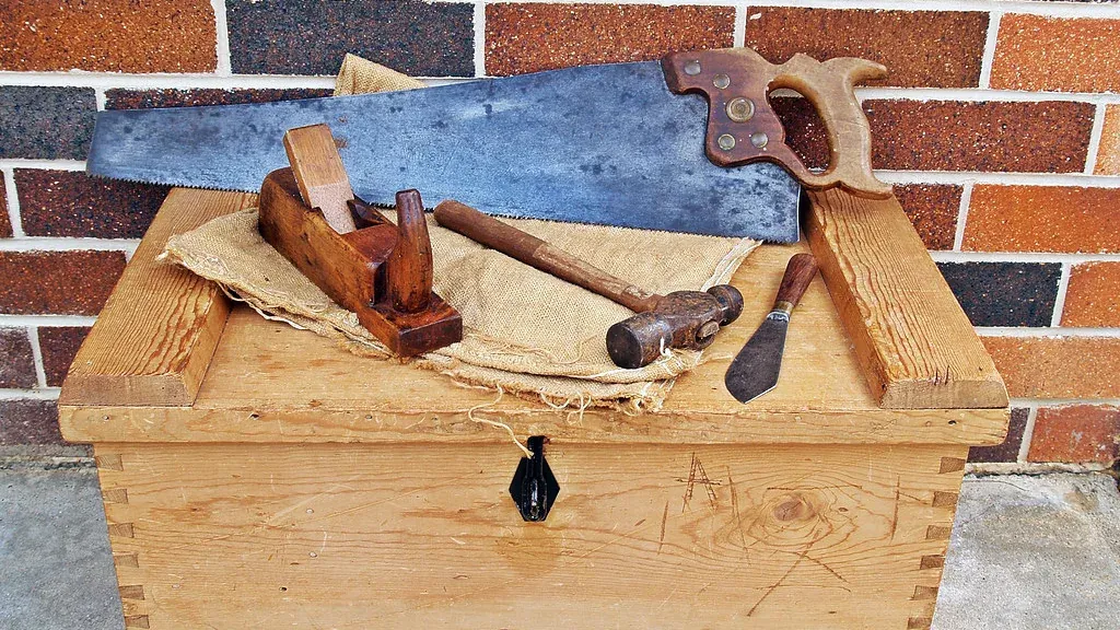 Hand saw, wooden plane, hammer and chisel laid on a wooden tool chest against a brick wall